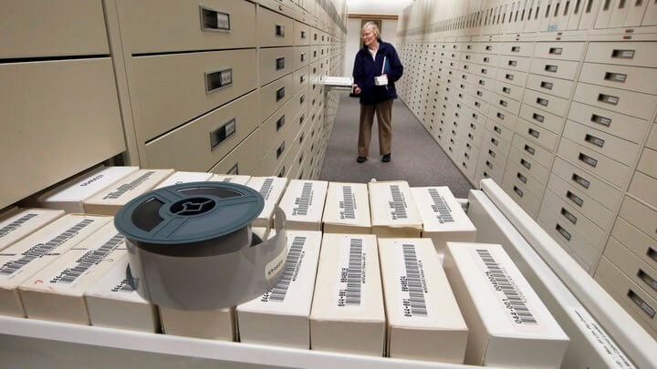 Individual standing in-between file drawers full of microfilm and microfiche. Image is focused on one open drawer filled with boxes of microfilm with a roll of microfilm placed on top.