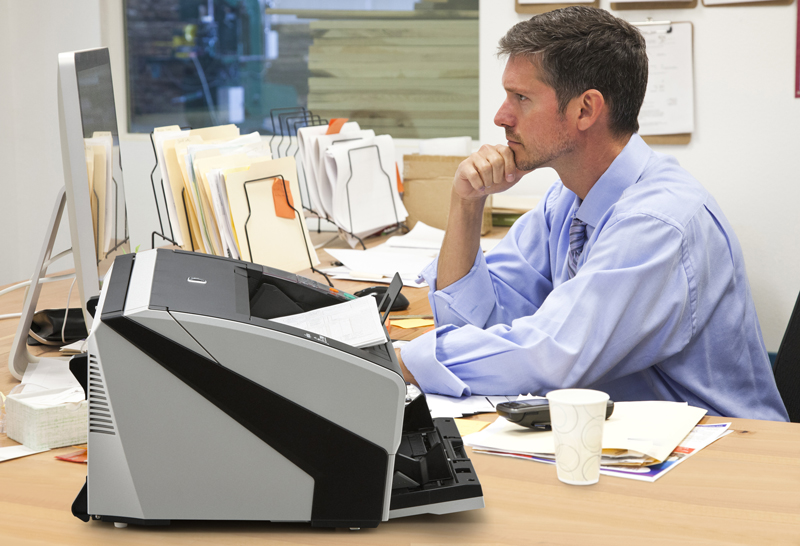 Low Profile Design Man sitting at desk looking at a computer screen with paper files and low profile scanner on desk. Image is meant to highlight how the scanner is low profile enough to sit on a desktop