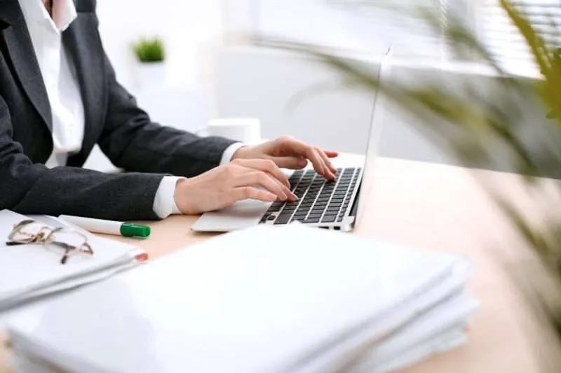 image of a woman's arms and hands typing case management notes on a laptop next to a stack of papers on desk